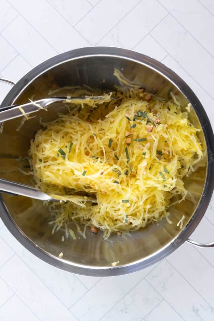 Stainless steel bowl containing cooked spaghetti squash strands mixed with herbs and nuts, with metal tongs resting inside.