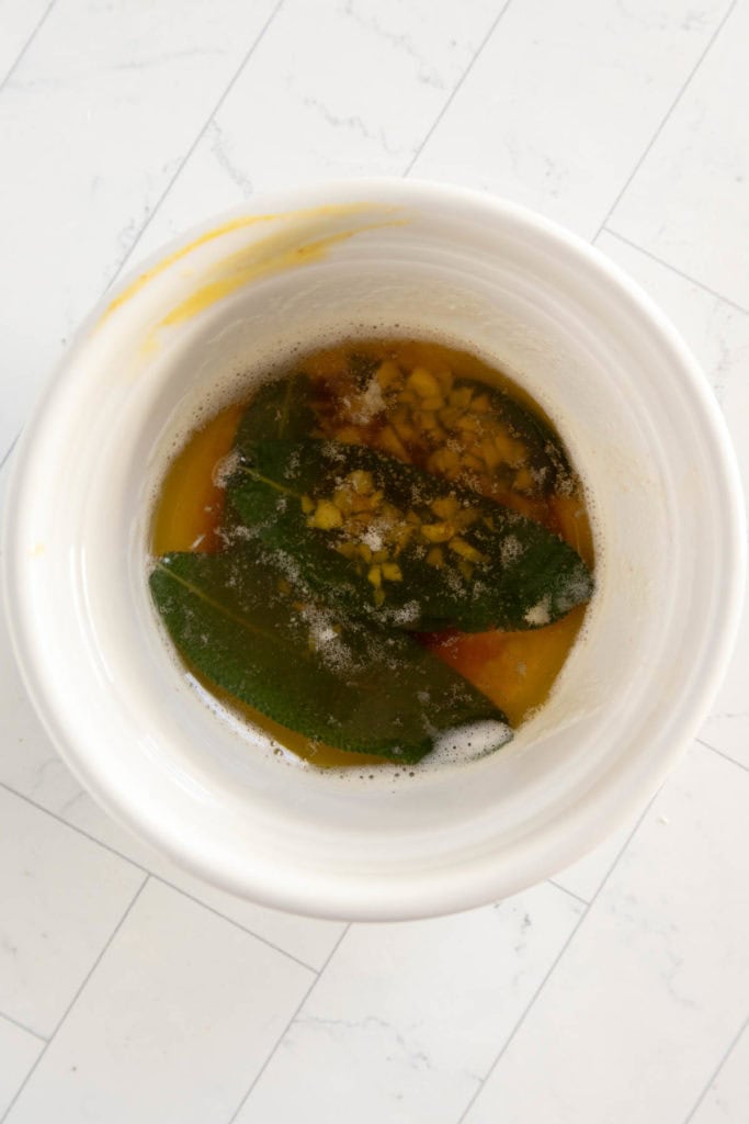 A white bowl containing melted butter, fresh sage leaves, and minced garlic, placed on a light-colored tiled surface.
