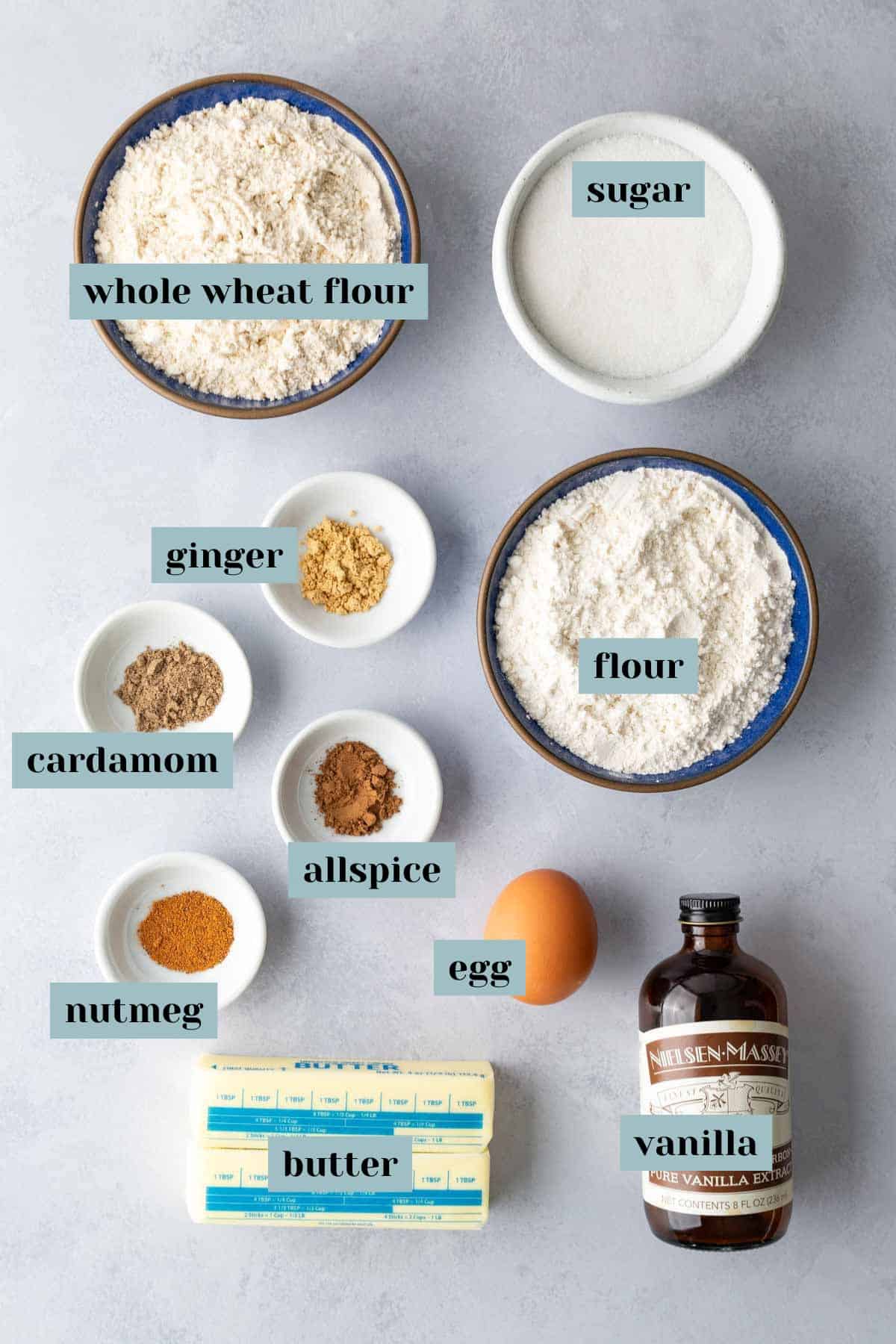 Bowls and plates of whole wheat flour, sugar, flour, ginger, cardamom, allspice, nutmeg, egg, butter sticks, and a bottle of vanilla on a light surface.