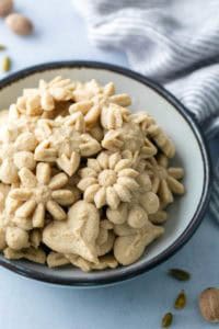 A bowl filled with assorted flower- and heart-shaped pressed cookies, with a striped cloth and whole spices nearby.