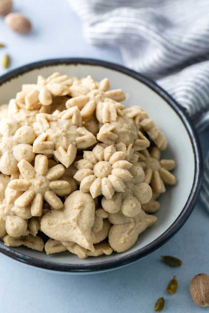 Spiced Spritz Cookies A bowl filled with assorted flower- and heart-shaped pressed cookies, with a striped cloth and whole spices nearby.