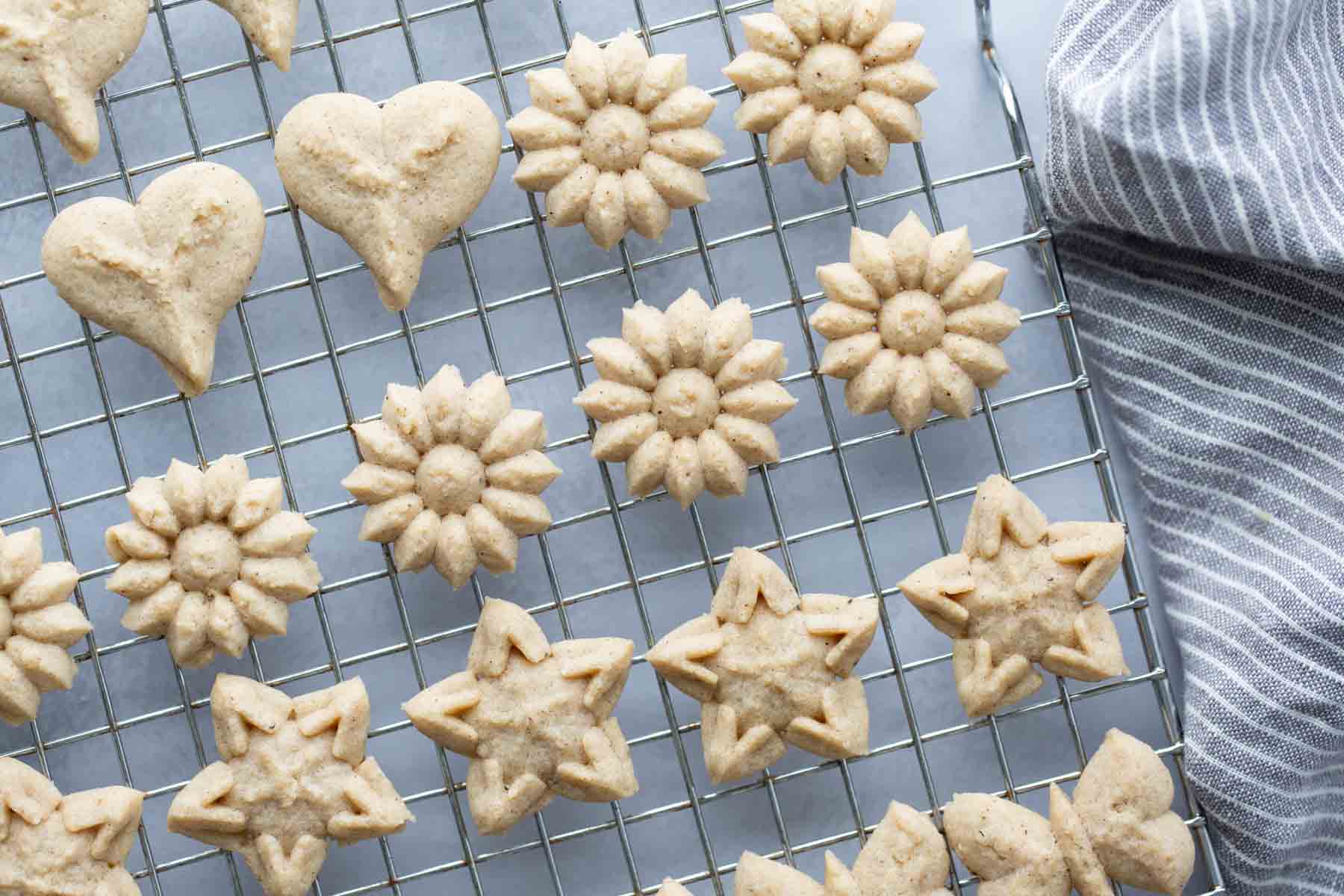 Assorted shaped cookies, including hearts, flowers, and stars, arranged on a cooling rack next to a striped cloth.