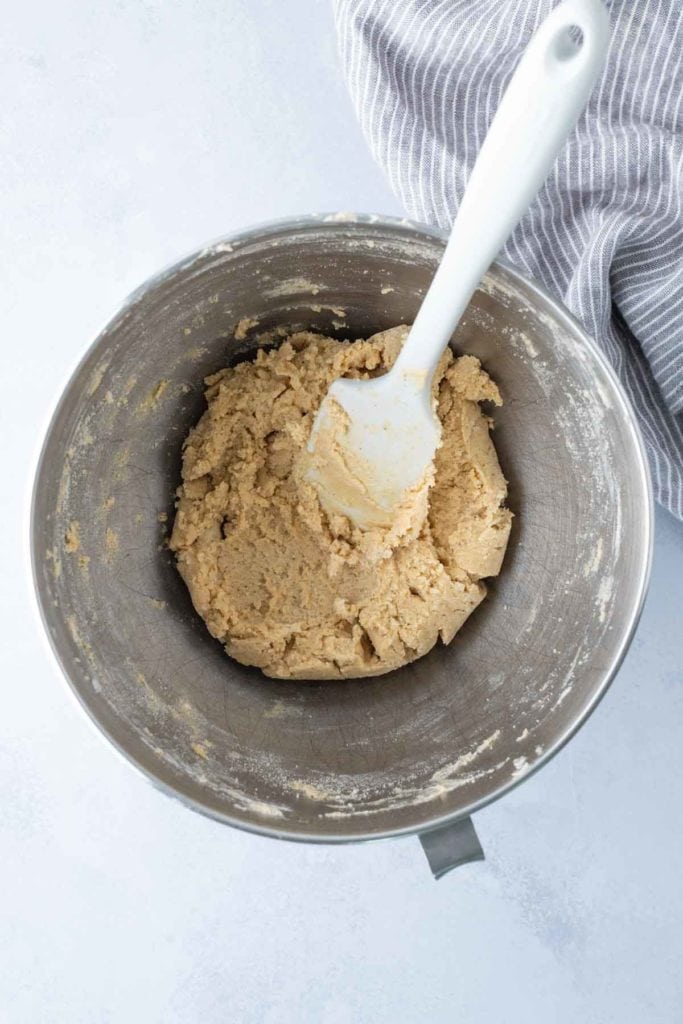 A metal mixing bowl with cookie dough and a white spatula, placed on a light surface next to a striped cloth.