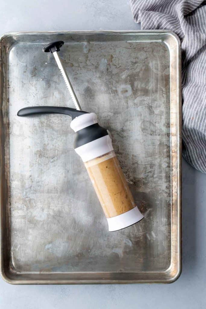 A cookie press filled with dough rests on a metal baking sheet next to a striped kitchen towel.