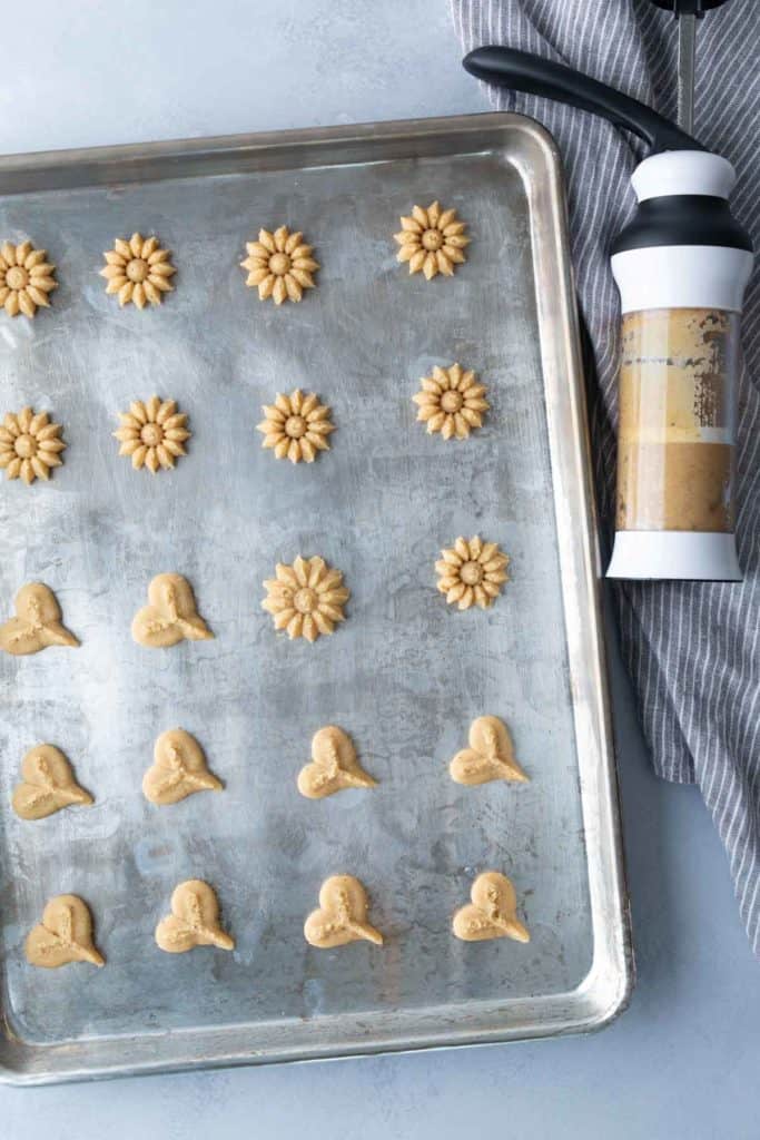 Unbaked flower and heart-shaped cookies arranged on a metal baking sheet, with a cookie press and striped cloth placed to the right.