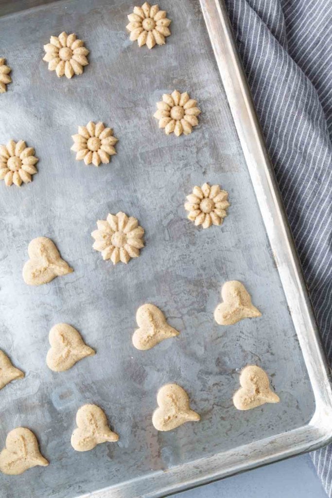 A metal baking sheet with unbaked flower and heart-shaped cookie dough pieces arranged in rows, next to a gray striped cloth.