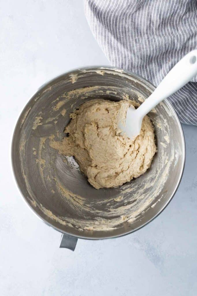 A stainless steel mixing bowl containing cookie dough and a white spatula, placed on a light surface beside a striped cloth.