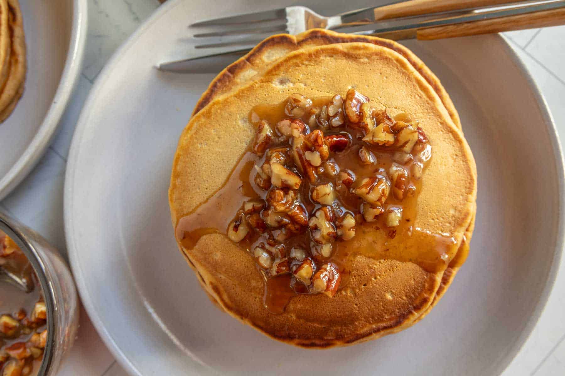 A stack of pancakes topped with chopped walnuts and syrup on a white plate, with a fork and knife placed nearby.