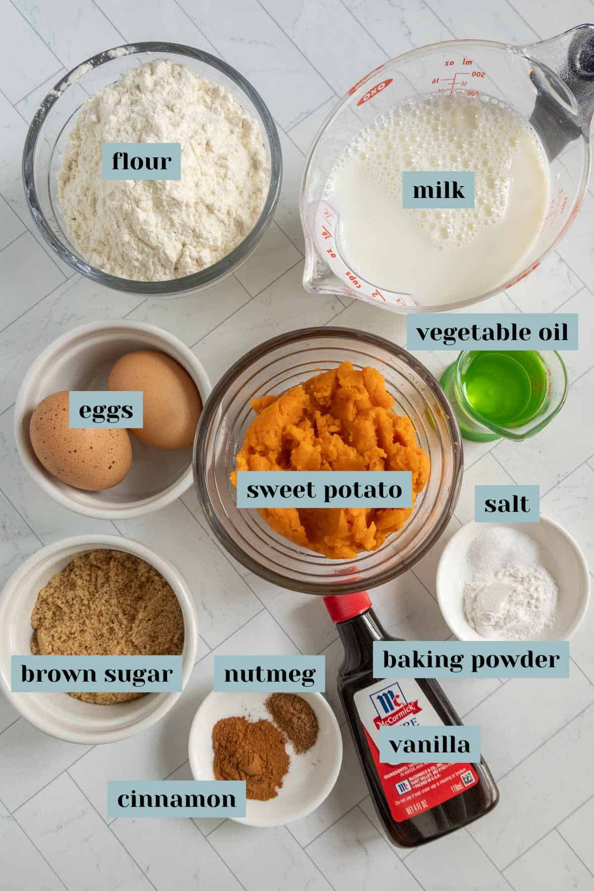 Top-down view of labeled ingredients for baking: flour, milk, vegetable oil, eggs, sweet potato, salt, brown sugar, nutmeg, baking powder, vanilla, and cinnamon.