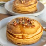 A plate of thick pancakes topped with syrup and chopped pecans, with another plate of pancakes in the background.