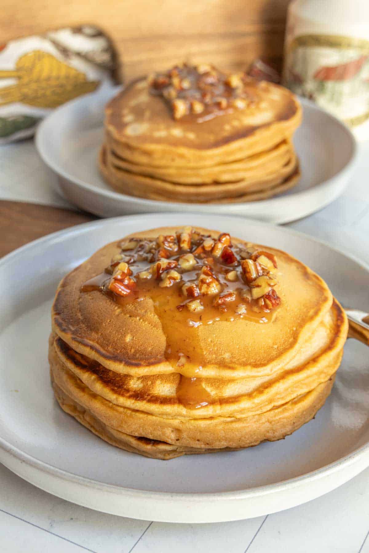 A plate of thick pancakes topped with syrup and chopped pecans, with another plate of pancakes in the background.
