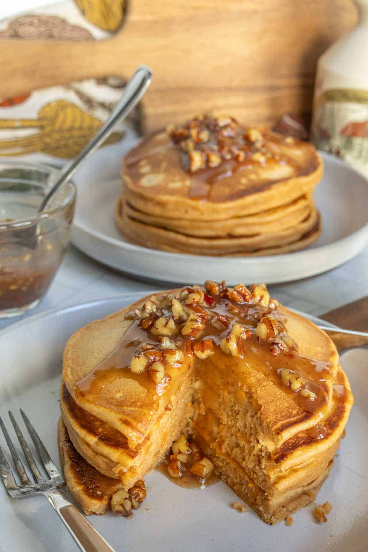 Stack of pancakes topped with chopped nuts and syrup, with a wedge cut out to show the inside. Another plate of pancakes and a glass jar are in the background.