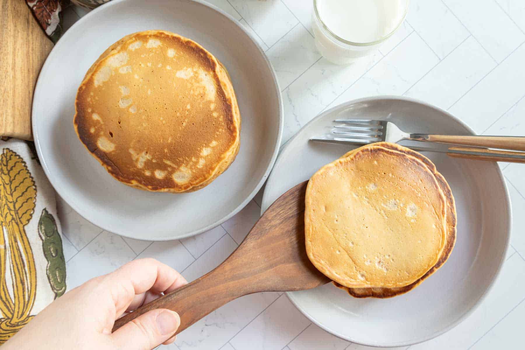 A hand uses a wooden spatula to place a pancake on a plate, next to another plate with a pancake, fork, knife, and a glass of milk on a white tiled surface.