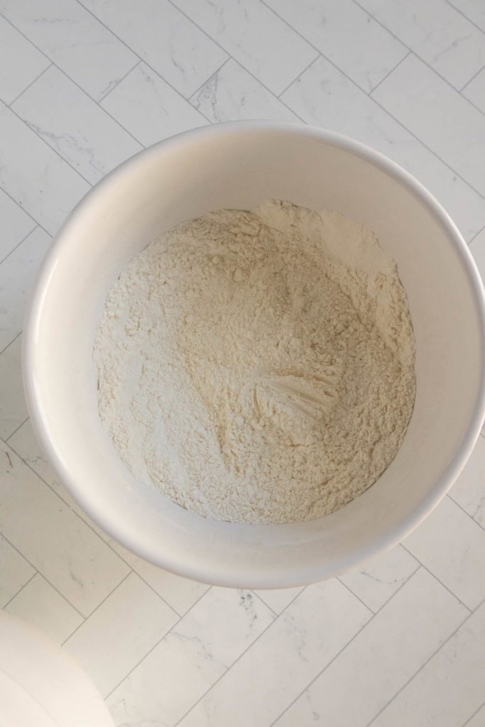A white mixing bowl filled with dry flour sits on a white tiled countertop.