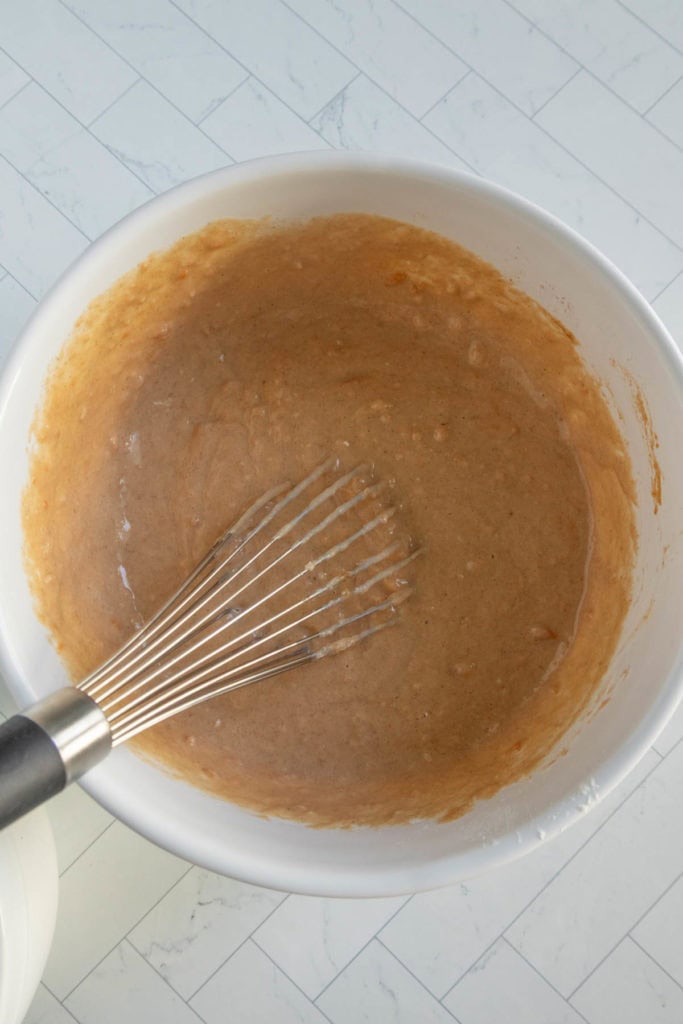 A metal whisk in a white bowl containing light brown batter sits on a white tiled surface.