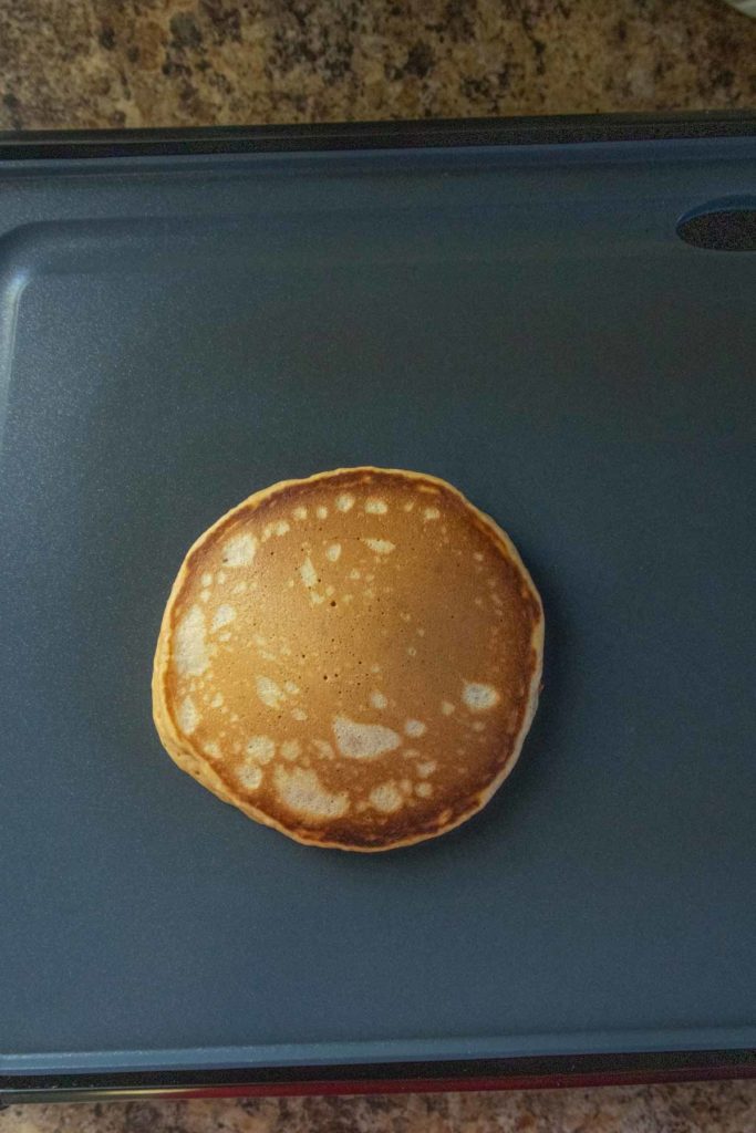 A single golden-brown pancake sits on a blue nonstick griddle, with a speckled countertop in the background.