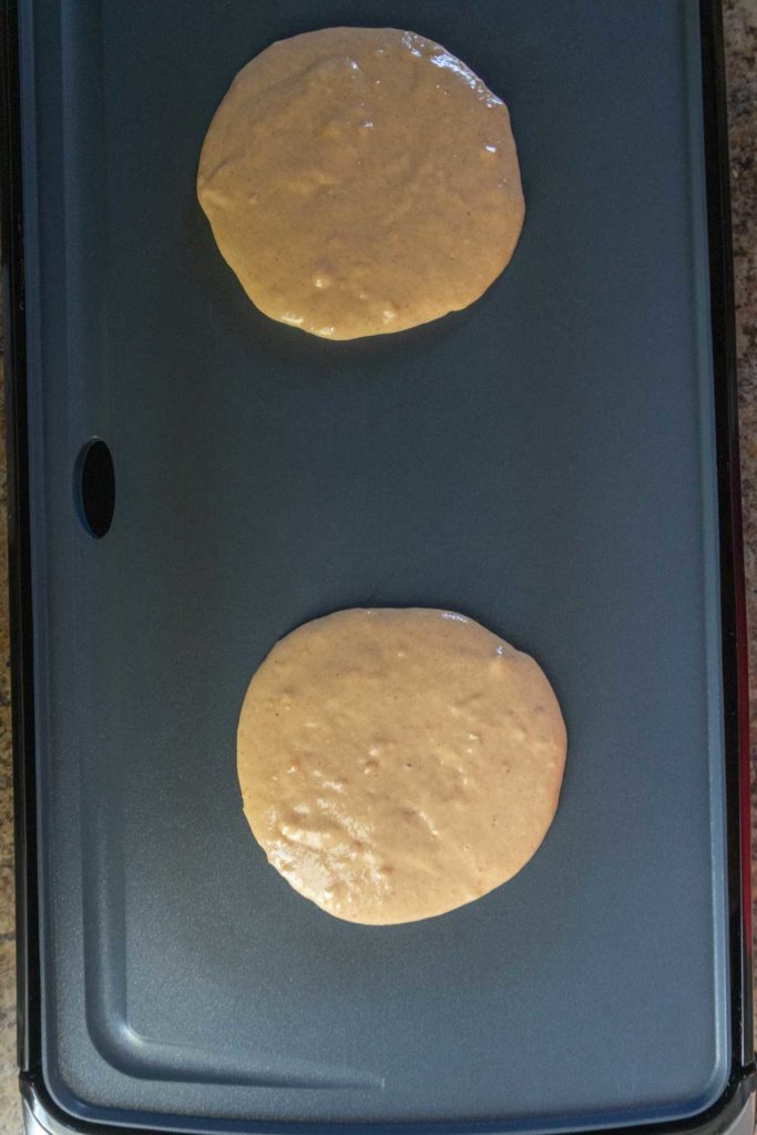 Two circles of pancake batter cooking on a flat, nonstick electric griddle.