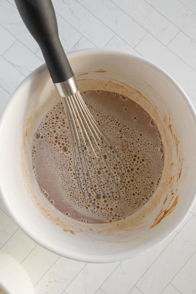 A whisk in a white bowl containing a brown liquid mixture with some bubbles on the surface, placed on a light-colored countertop.