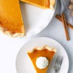 A pumpkin pie with one slice removed sits on a plate; the slice is served on a separate plate topped with whipped cream and a fork beside it. Cinnamon sticks are nearby.