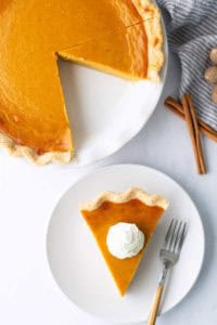 A pumpkin pie with one slice removed sits on a plate; the slice is served on a separate plate topped with whipped cream and a fork beside it. Cinnamon sticks are nearby.