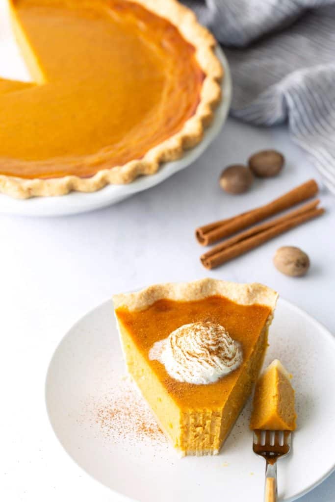 15 Craveworthy Thanksgiving Pies for a Sweet Finish A slice of pumpkin pie with whipped cream on a white plate, next to a forkful of pie; whole pie, cinnamon sticks, and nutmeg in the background.