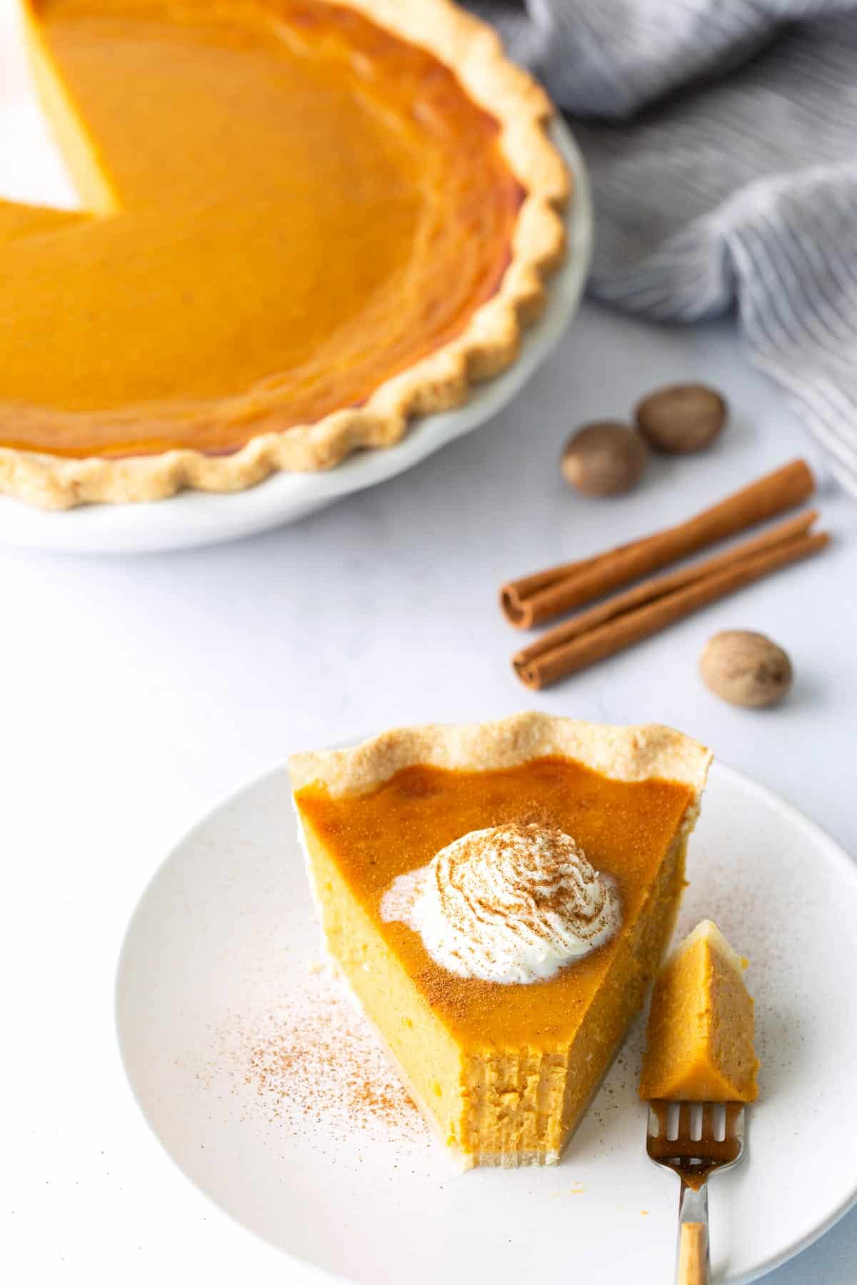 A slice of pumpkin pie with whipped cream on a white plate, next to a forkful of pie; whole pie, cinnamon sticks, and nutmeg in the background.