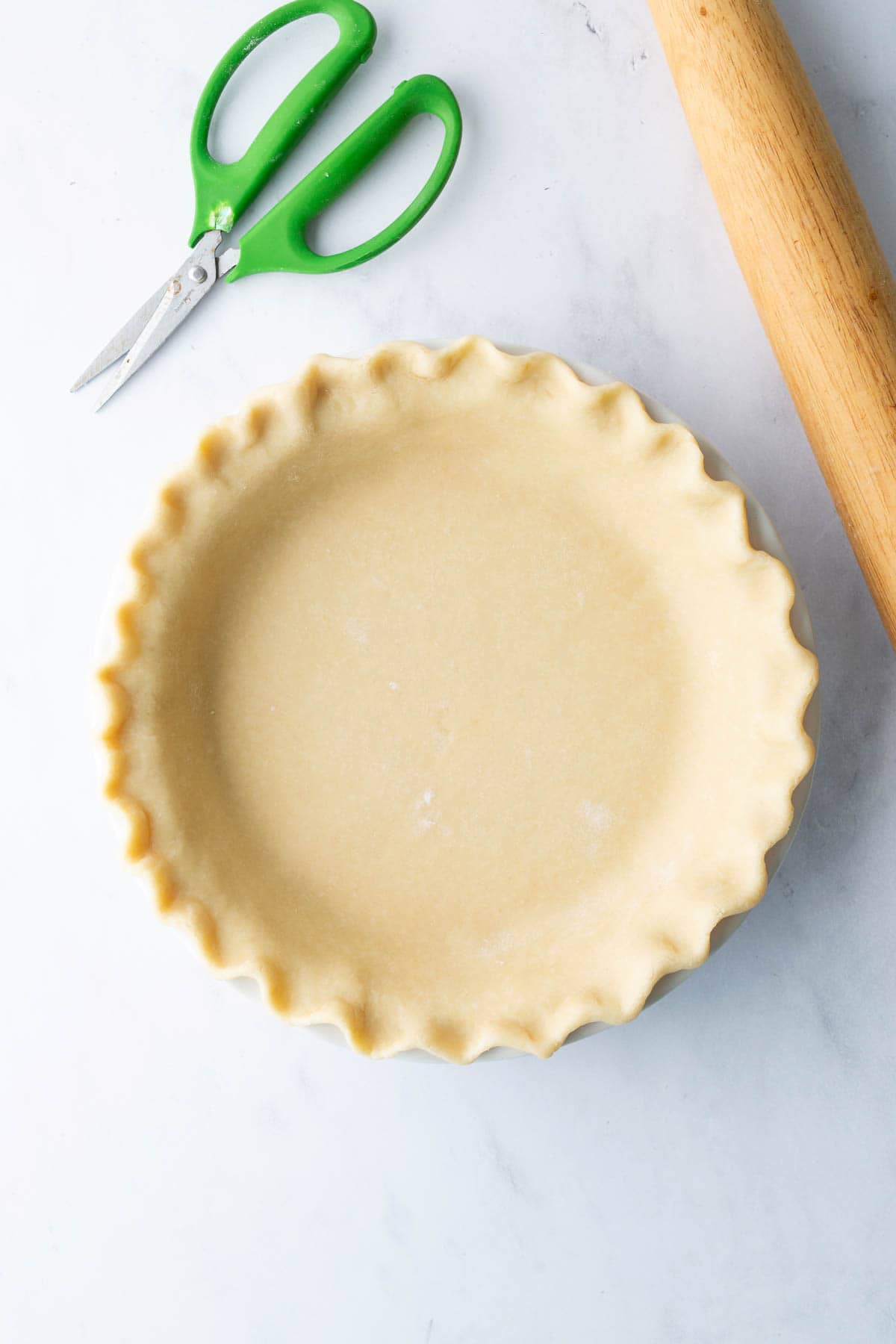 Unbaked pie crust in a fluted pie dish on a white surface, with green-handled scissors and a wooden rolling pin nearby.