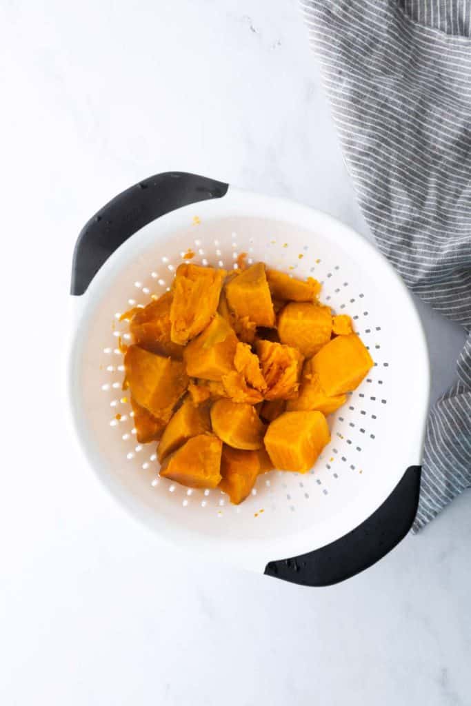 Chunks of cooked sweet potato sit in a white plastic colander with black handles, next to a gray striped cloth on a white surface.
