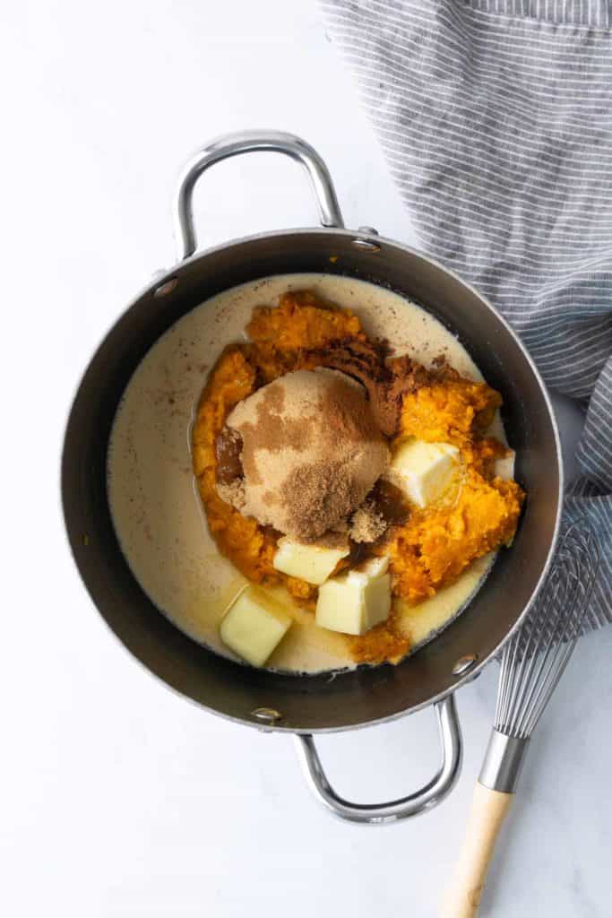 A saucepan with mashed sweet potatoes, brown sugar, butter, and cream, placed beside a striped towel and a whisk on a white surface.