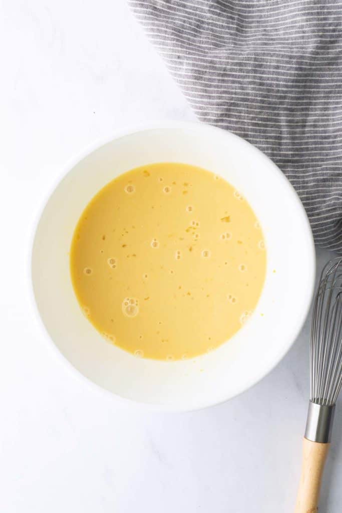 A white bowl with a light yellow liquid mixture, possibly batter, sits on a white surface next to a wire whisk and a gray-striped cloth.