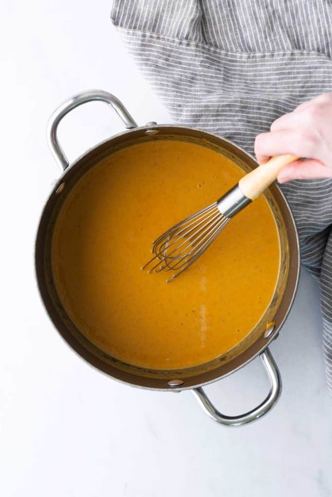 A hand uses a whisk to stir orange-colored sauce in a metal pot, with a gray-striped cloth partially visible beside the pot.