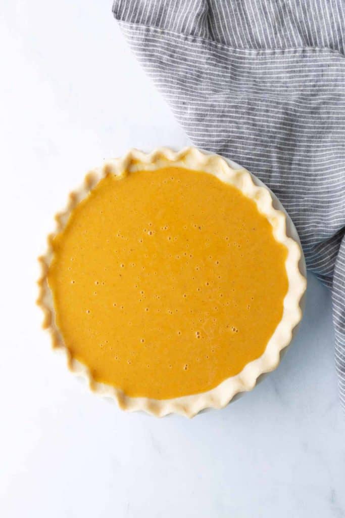 Unbaked pumpkin pie filling in a pie crust sits on a white surface next to a gray and white striped cloth.