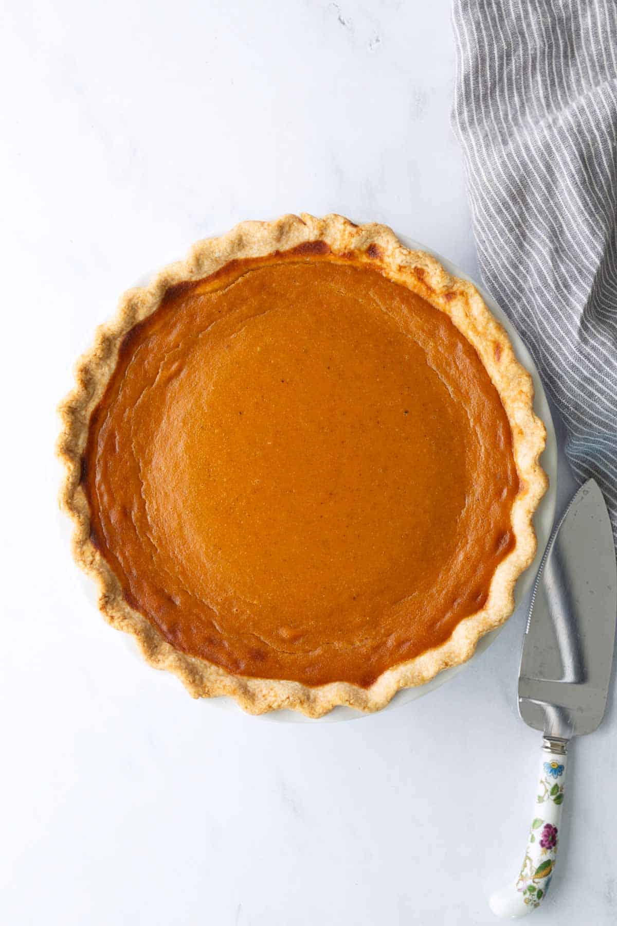 A whole baked pumpkin pie in a pie dish next to a pie server on a white marble surface with a gray striped cloth.