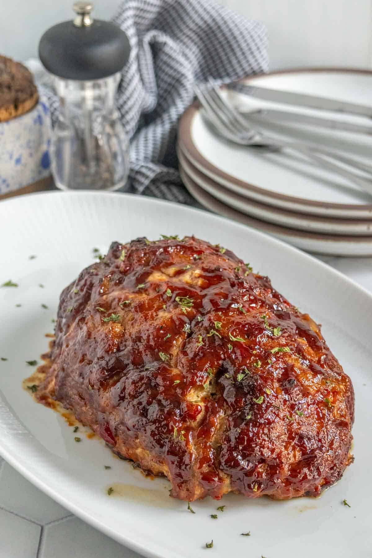 A glazed meatloaf garnished with herbs is served on a white platter, with stacked plates, utensils, and a pepper grinder in the background.