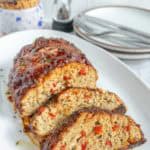 Sliced meatloaf with red pepper pieces on a white platter, served with plates and utensils in the background.