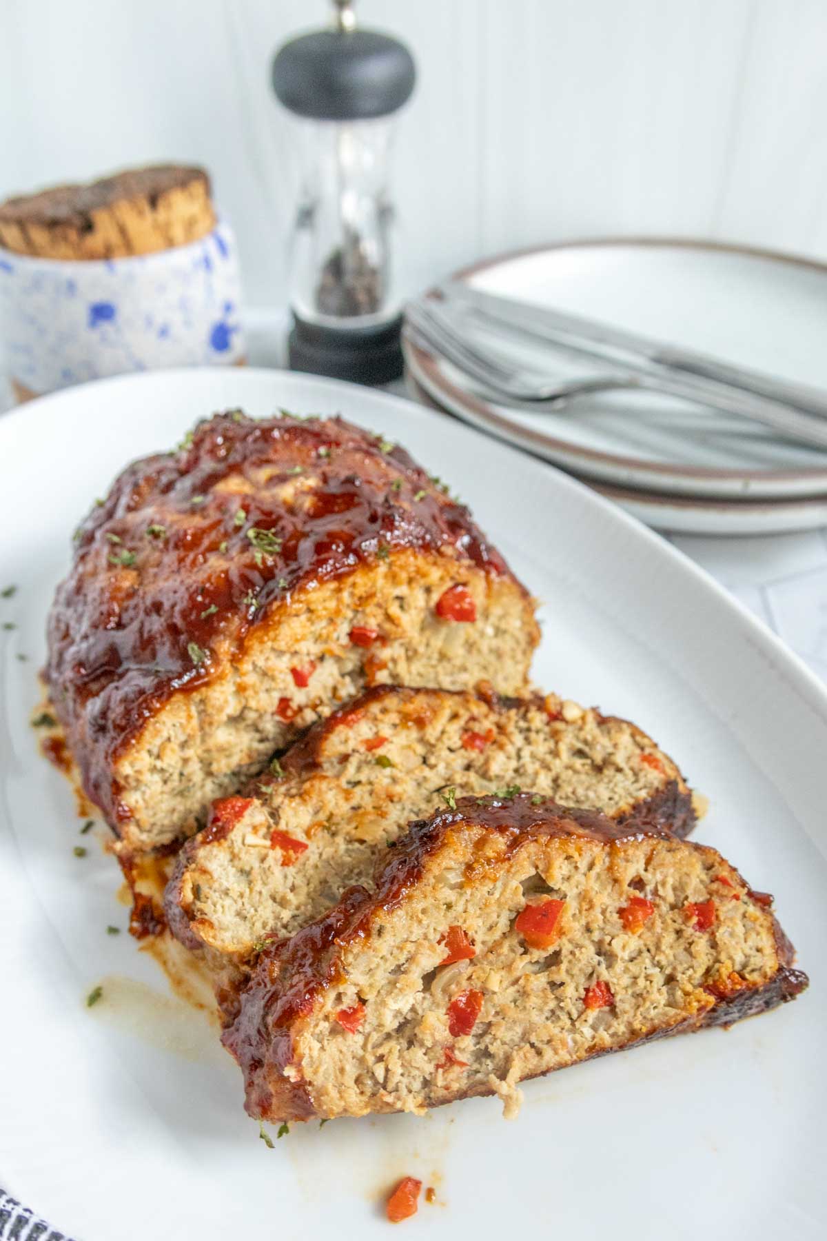 Sliced meatloaf with red pepper pieces on a white platter, served with plates and utensils in the background.