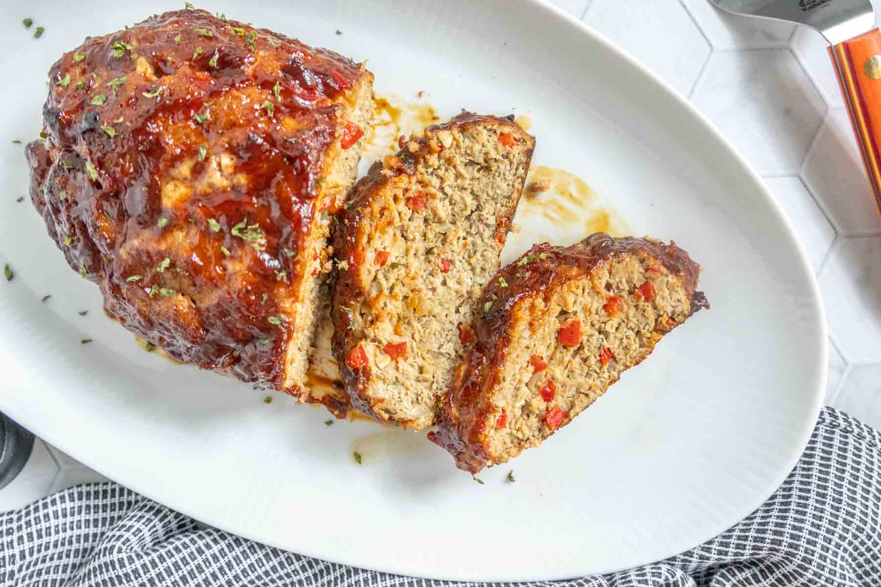 A sliced meatloaf with a glazed topping and visible red bell peppers inside, served on a white oval plate.