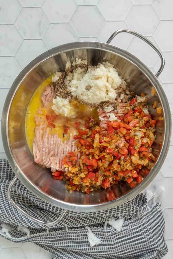 A metal mixing bowl with ground meat, sautรฉed vegetables, beaten eggs, breadcrumbs, and seasoning, placed on a black and white towel on a tiled surface.