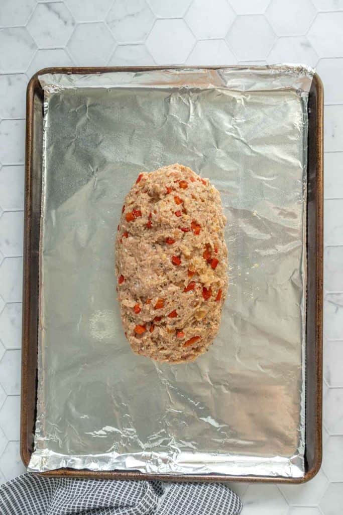 Uncooked meatloaf mixture with chopped red bell peppers shaped into a loaf on a foil-lined baking sheet.