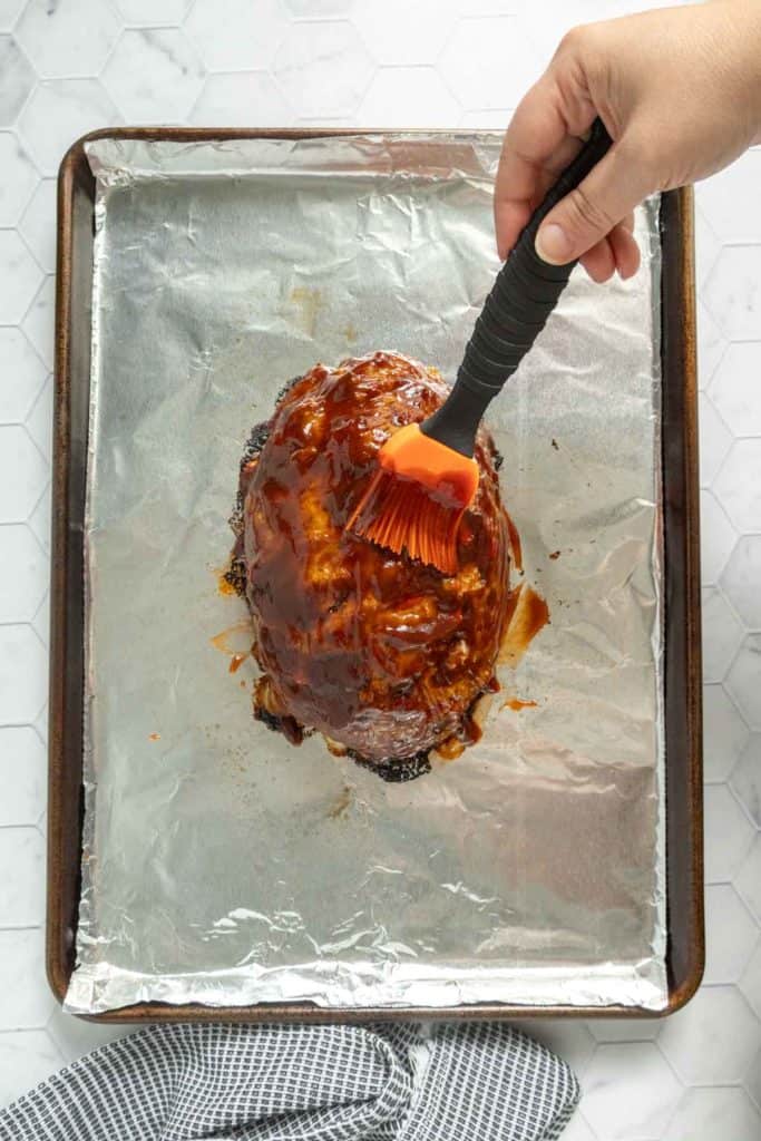 A hand uses a brush to spread sauce over a meatloaf on a foil-lined baking sheet, with a striped towel nearby.
