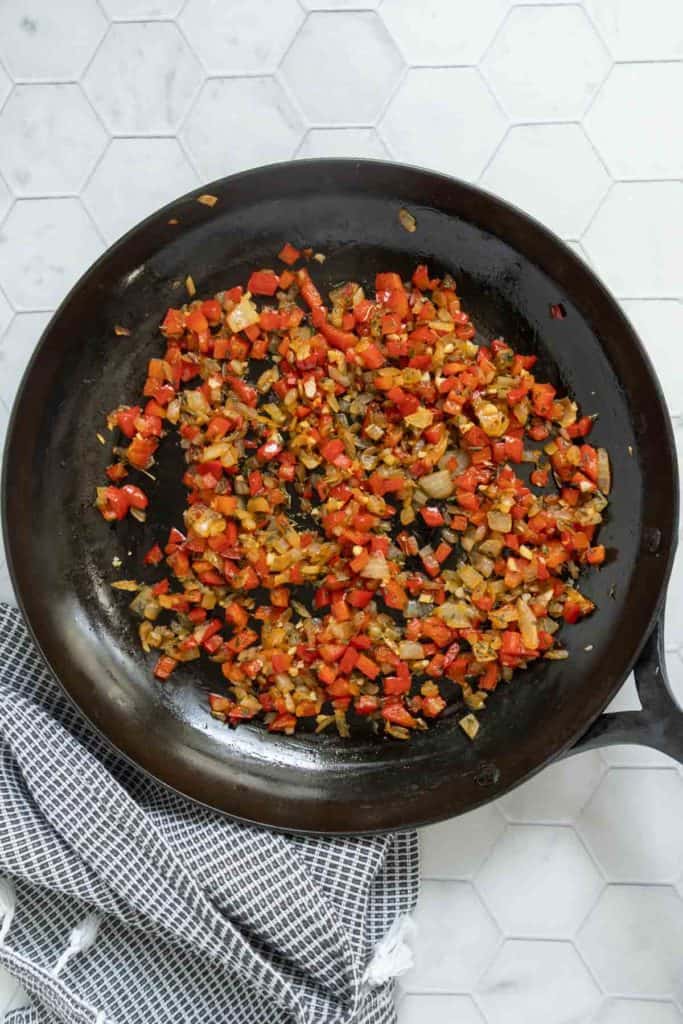 A frying pan on a stovetop contains sautรฉed diced red bell peppers and onions, with a checkered kitchen towel placed nearby.