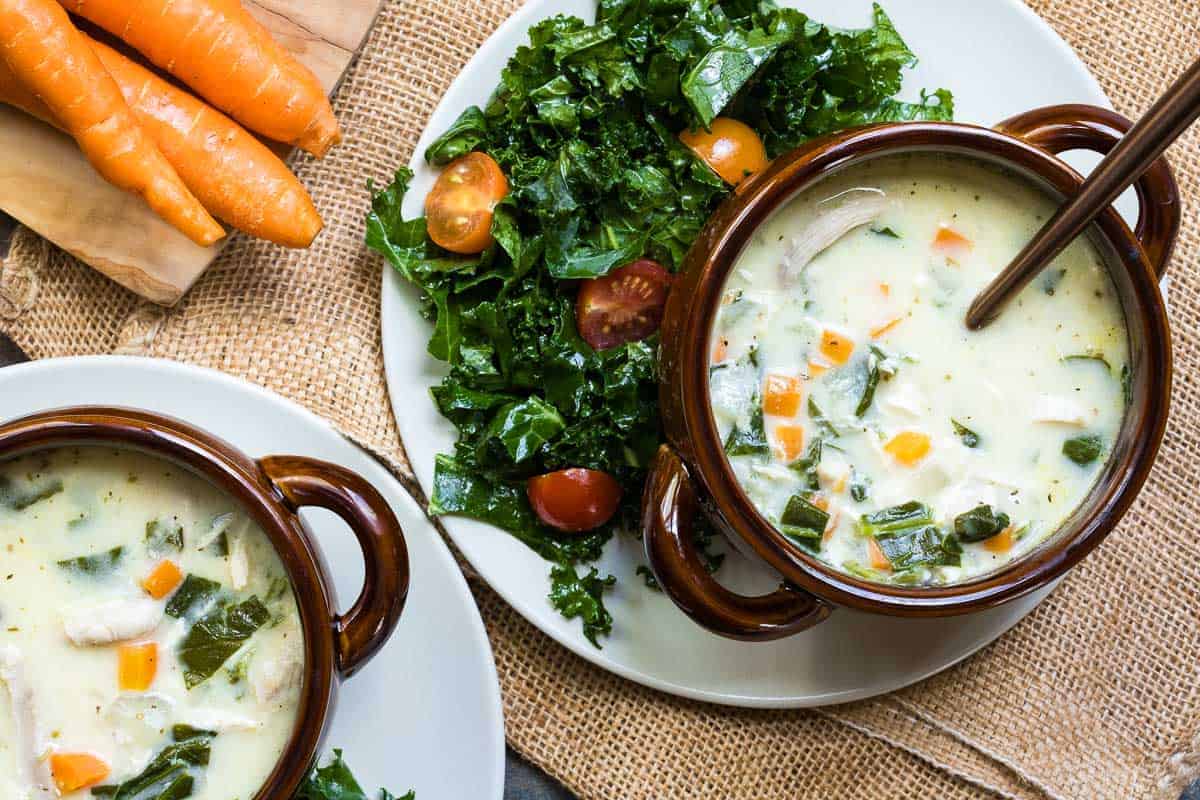 Two bowls of creamy vegetable and chicken soup are served alongside a fresh kale and tomato salad; raw carrots are placed nearby on a wooden board.