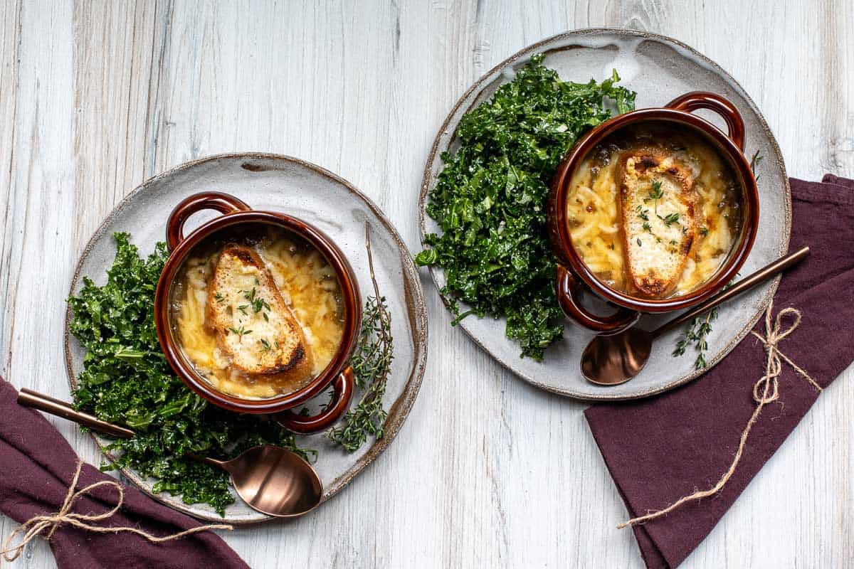 Two bowls of French onion soup topped with toasted bread and melted cheese, served on plates with a side of crispy kale and bronze-colored spoons, placed on a light wooden surface.