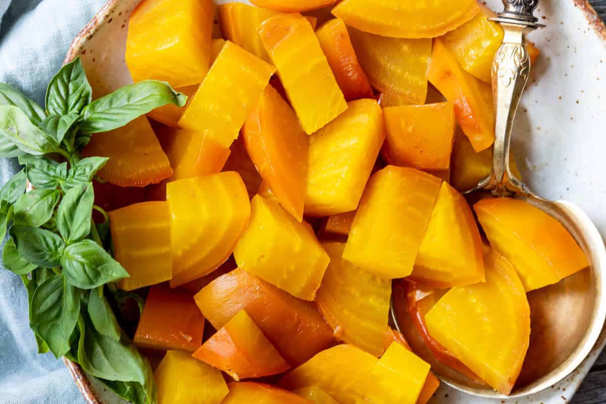 A plate of sliced cooked golden beets with a sprig of fresh basil and a metal serving spoon.