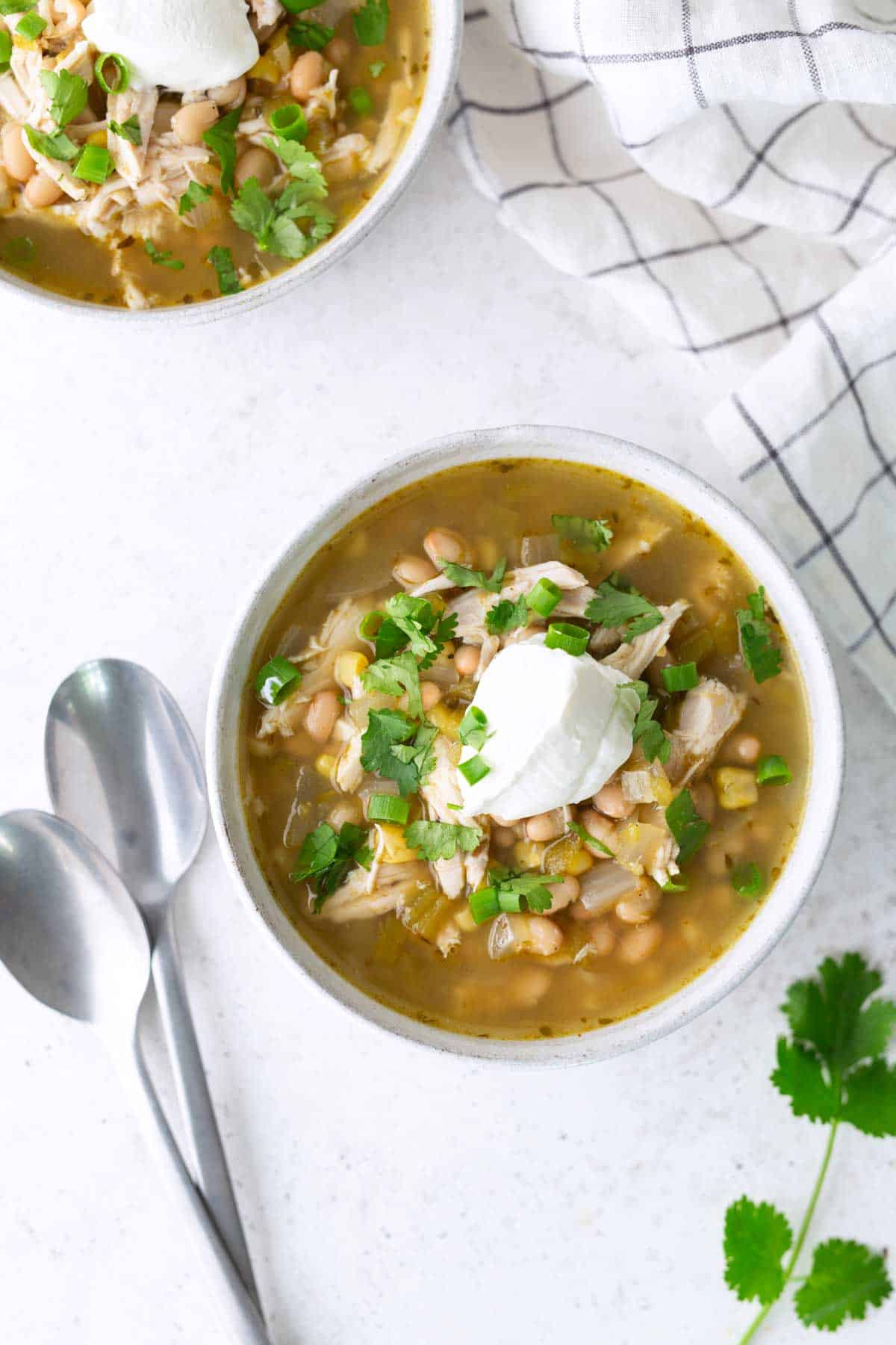 A bowl of white bean chicken chili topped with sour cream and chopped cilantro, next to two spoons and a cloth napkin.