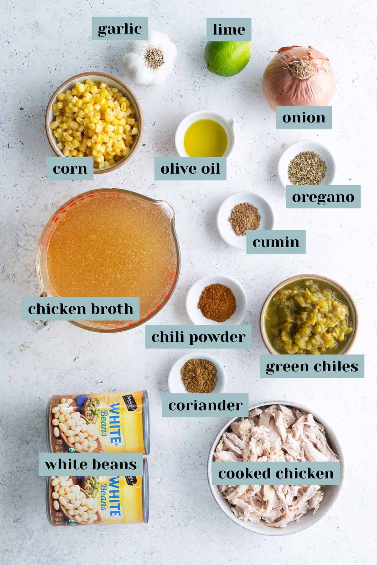 Ingredients for white chicken chili are arranged on a white surface, each labeled: corn, chicken broth, white beans, cooked chicken, garlic, lime, olive oil, onion, oregano, cumin, chili powder, coriander, green chiles.