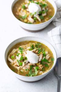 Two bowls of white chicken chili topped with sour cream, chopped green onions, and cilantro sit on a white surface next to a spoon and a checkered napkin.