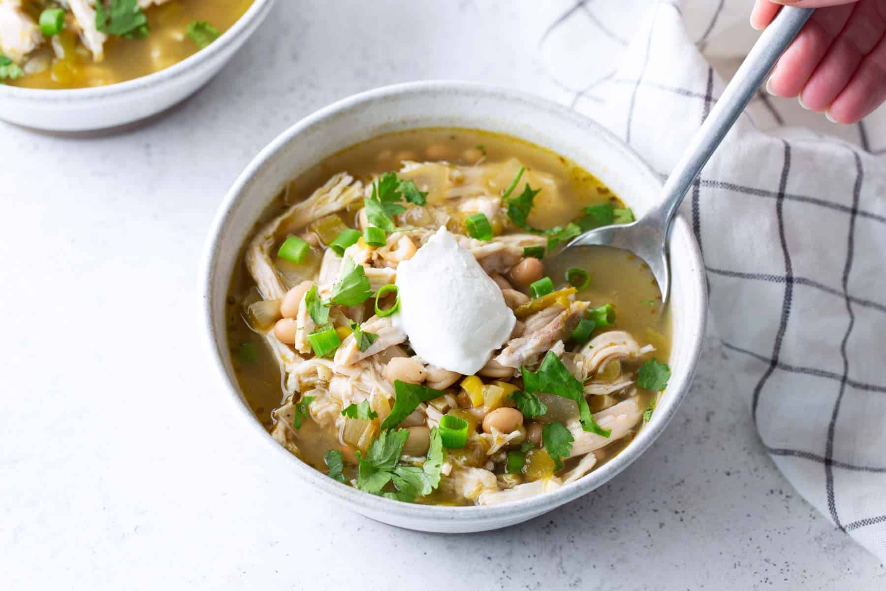 A bowl of white chicken chili topped with sour cream, chopped cilantro, and green onions, with a spoon and a checkered napkin beside it.