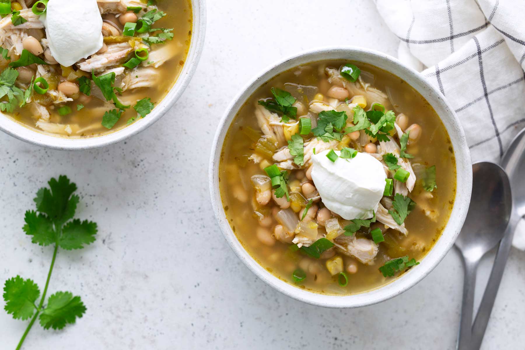 Two bowls of white chicken chili topped with sour cream and chopped herbs, placed on a light surface with a spoon, napkin, and cilantro sprig nearby.