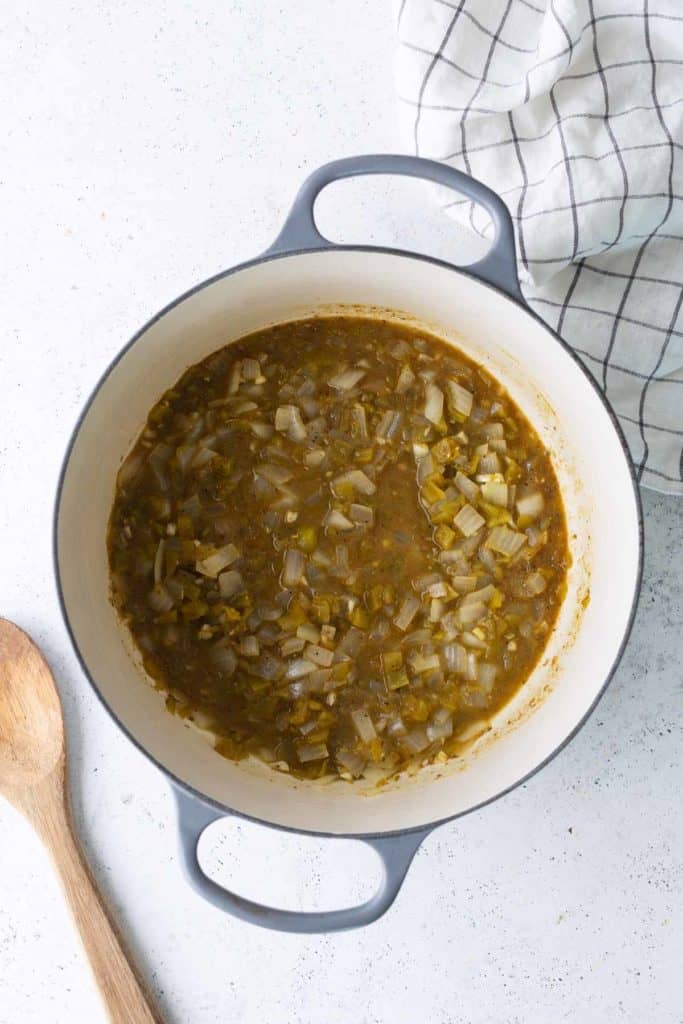 A Dutch oven with cooked diced onions and green chilies on a white surface, next to a wooden spoon and a checkered kitchen towel.